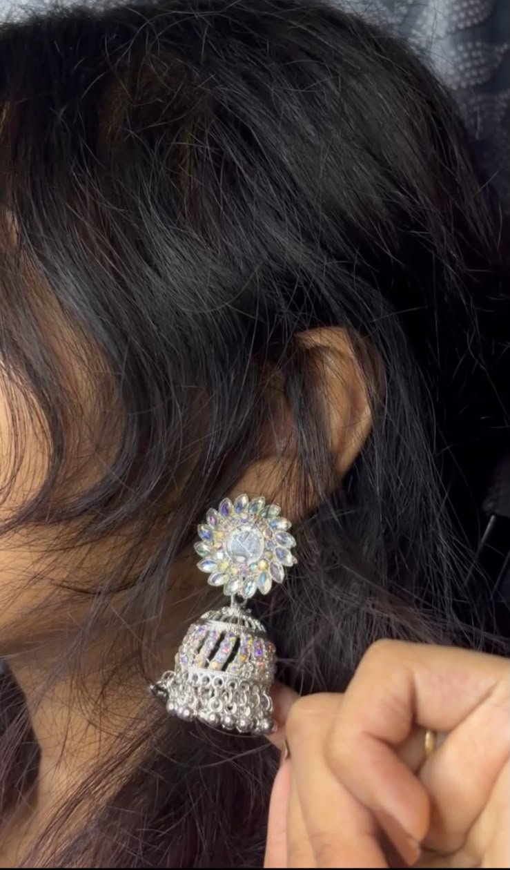 A close-up of a person wearing a large, ornate silver-toned jhumka earring adorned with clear stones, with dark hair and a hand adjusting the earring.