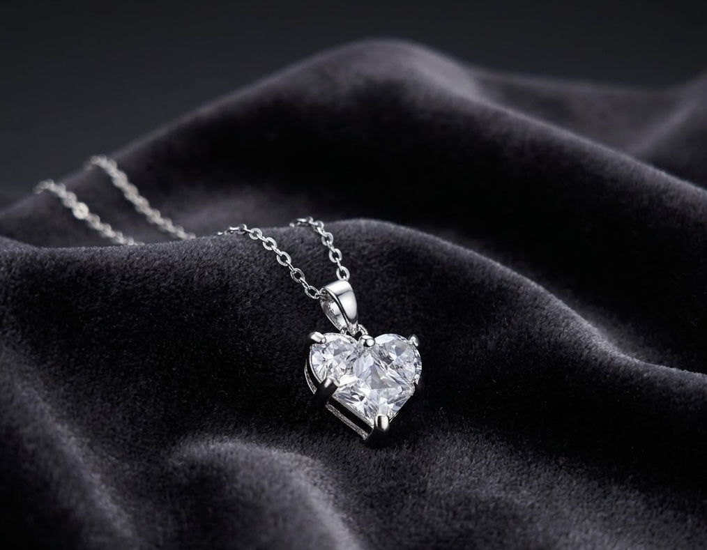 A silver heart-shaped pendant necklace with a clear faceted gemstone, resting on draped black velvet fabric.