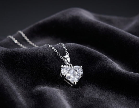 A silver heart-shaped pendant necklace with a clear faceted gemstone, resting on draped black velvet fabric.