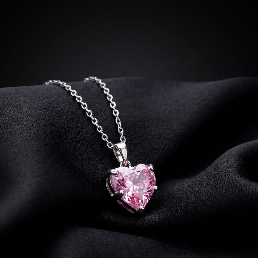 A silver heart-shaped pendant necklace with a sparkling pink gemstone, resting on draped black fabric.