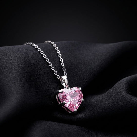 A silver heart-shaped pendant necklace with a sparkling pink gemstone, resting on draped black fabric.