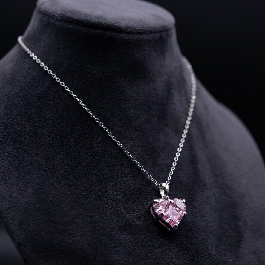 A silver heart-shaped pendant necklace with a pink gemstone, displayed on a black velvet jewelry bust against a dark background.