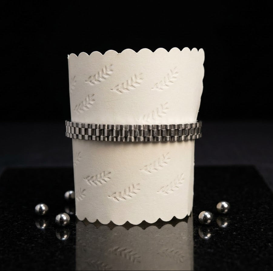 A white paper cup sleeve with scalloped edges and an embossed leaf pattern, wrapped with a silver metallic beaded bracelet, displayed on a black reflective surface with small silver beads.