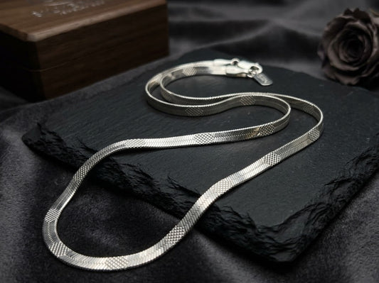 A silver snake chain necklace with a textured mesh pattern, draped over a black slate tray on a black satin fabric background, with a dark rose and a wooden box in the background.