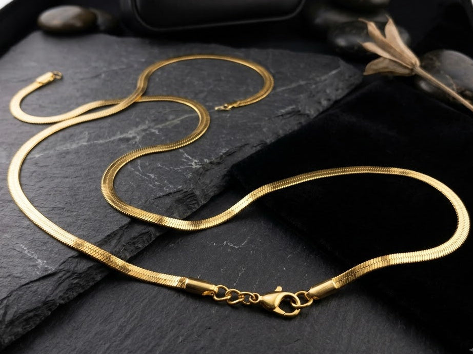 A gold snake chain necklace displayed on a black velvet surface with a black slate slab, small dark stones, and a black Aurora Goldsmith box in the background.