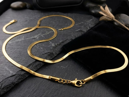 A gold snake chain necklace displayed on a black velvet surface with a black slate slab, small dark stones, and a black Aurora Goldsmith box in the background.