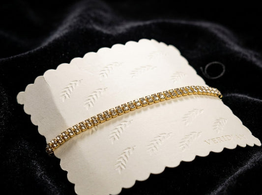 A gold tennis bracelet with clear stones, displayed on a white scalloped card over a black velvet background.