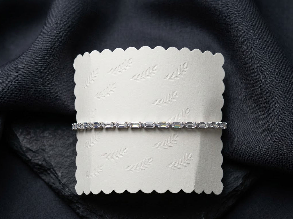 A silver-toned diamond tennis bracelet displayed on a white scalloped paper card, set against a dark black fabric background.
