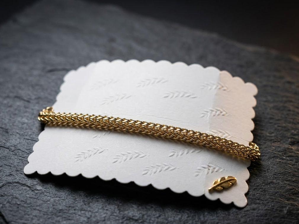 A gold chain bracelet with a small gold leaf charm, displayed on a white scalloped paper card over a dark slate surface.